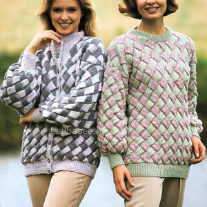 May include: Two women wearing knitted sweaters with a woven basket weave design. One sweater is a button-up cardigan in shades of gray and white. The other is a crew neck sweater in shades of pink and green. Both sweaters have long sleeves.