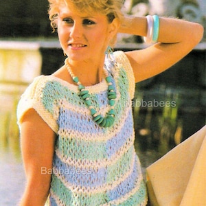 Op de afbeelding: Een gehaakte top in blauwe, groene en crèmekleurige tinten, met een chevronpatroon. De top heeft korte mouwen en een ronde halslijn. De afbeelding toont ook een turquoise ketting en bijpassende oorbellen.