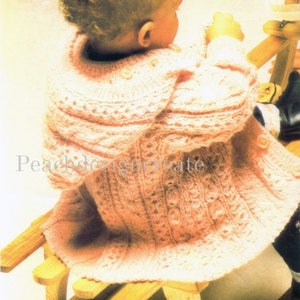 May include: A pink knitted baby cardigan with a cable knit pattern and a hood. The cardigan is worn by a baby sitting on a wooden rocking horse.