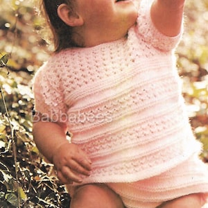 May include: A baby wearing a pink crocheted dress with a lacy pattern. The baby is sitting in a grassy area with green leaves.