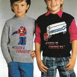 May include: Two boys wearing knitted sweaters with Transformers designs. The boy on the left is wearing a gray sweater with a red and blue robot and the text "The Transformers". The boy on the right is wearing a black sweater with a white truck and the text "The Transformers".