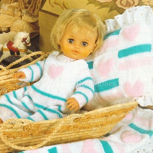 Puede incluir: Una muñeca con un conjunto de punto blanco con un corazón rosa y rayas turquesas, en una cesta de mimbre. También hay una almohada y una manta a juego con corazones rosas y rayas turquesas.