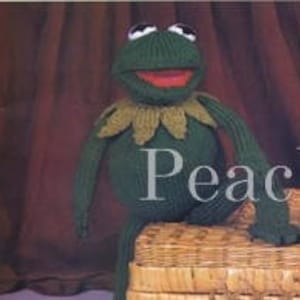 May include: Green knitted frog puppet with a large mouth and googly eyes. The frog is wearing a yellow knitted collar.