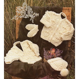 May include: A set of knitted baby clothes in a cream color, including a cardigan, a hat, and mittens. The clothes are laid out on a wicker basket with a plaid fabric background.