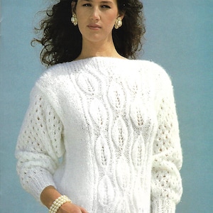 May include: A white knitted sweater with a cable knit pattern and a delicate lace design. The sweater has a boat neck and long sleeves.