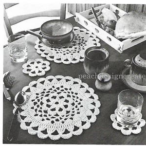 Puede incluir: Un juego de cuatro manteles individuales de ganchillo en color blanco. Los manteles individuales son redondos y tienen un delicado diseño floral. Los manteles individuales están dispuestos sobre una mesa con una taza de café, un vaso de agua y un cuenco.