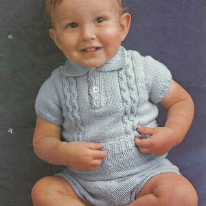 May include: A baby wearing a light blue knitted onesie with a cable knit pattern and buttons. The onesie has short sleeves and a collar.