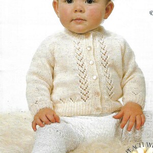 May include: A white knitted baby cardigan with a button-up front and a delicate lace pattern. The cardigan is worn by a baby sitting on a white fluffy surface.