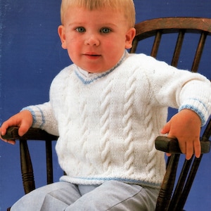May include: A white knitted sweater with a cable knit pattern and blue trim. The sweater has a V-neck and is worn by a young child sitting on a wooden chair.