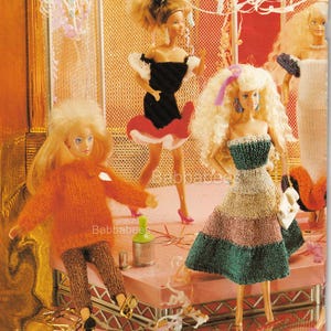 May include: Four dolls wearing knitted outfits. The dolls are standing on a stage with a pink platform and a white metal frame. The dolls are wearing a variety of outfits, including a black dress with a red skirt, a green and pink dress, a brown sweater and trousers, and a white dress with a purple trim.