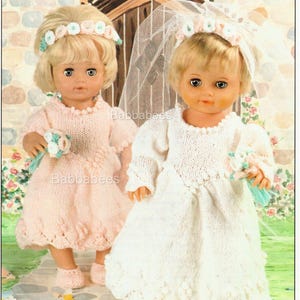 May include: Two dolls dressed in knitted outfits. One doll wears a pale pink dress, headband, and shoes, holding a small posy. The other doll is in a white wedding dress with a veil and floral headband, also holding a bouquet. The text "Babbabees" is visible.