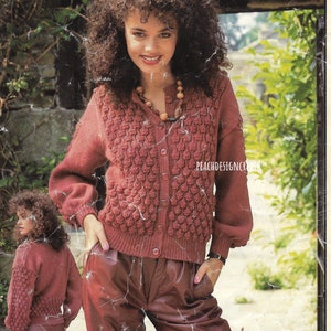 May include: A close-up of a woman wearing a pink knitted cardigan with a textured pattern and a button closure. She is wearing brown leather pants and a beaded necklace.