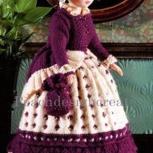 May include: A knitted doll wearing a purple dress with a white lace trim and a white bonnet. The doll has brown hair and blue eyes.