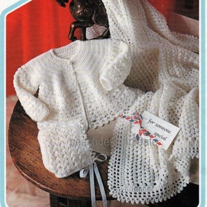 May include: A white crocheted baby set including a cardigan, bonnet, and blanket. The blanket has a square design and the cardigan has a button closure. The set is displayed on a wooden surface with a note that reads "...for someone special."