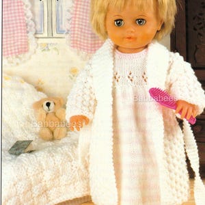 May include: A doll with blonde hair in a white knitted robe and nightdress, holding a pink brush. A small teddy bear and a book are on the bed. The scene is set in a bedroom with a window and curtains.