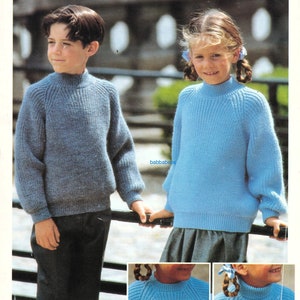 May include: Two children wearing blue knitted jumpers with mock necks. The jumpers have a ribbed texture. The boy is wearing blue jeans and the girl is wearing a grey skirt.
