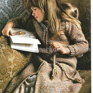 May include: A young girl wearing a brown and white knitted hooded cardigan with a zipper closure. She is sitting on a couch and reading a book. The cardigan has a tie belt and a fringed hem.