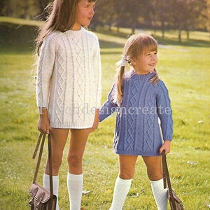 May include: Two young girls wearing white and blue knitted dresses with cable knit patterns. They are both wearing white socks and brown shoes. The girl on the left is holding a brown leather satchel.