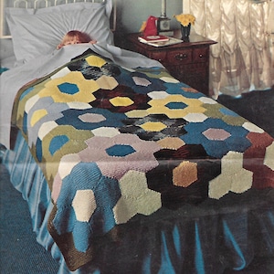 May include: A colourful knitted blanket with a geometric pattern of hexagons in shades of blue, green, yellow, brown, and purple. The blanket is on a bed with a blue bed skirt.