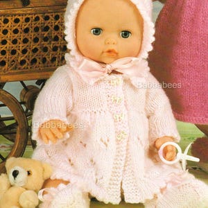 Può includere: Una bambola vestita con un completo in maglia rosa chiaro, tra cui cappello, cappotto e stivaletti. Il cappotto ha bottoni decorativi e un fiocco. Sono visibili anche un piccolo orsacchiotto e un giocattolo. La bambola è vicino a un passeggino vintage in legno.