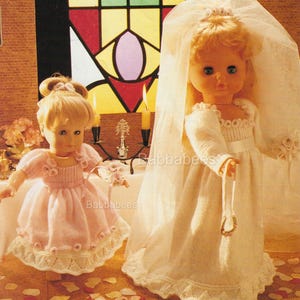 Puede incluir: Dos muñecas vestidas con vestidos de punto blanco y rosa. La muñeca de la izquierda lleva un vestido rosa con ribete de encaje blanco y una flor blanca en el hombro. La muñeca de la derecha lleva un vestido blanco con un velo blanco y una flor blanca en el hombro. Ambas muñecas sostienen flores blancas.