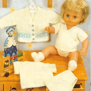 Op de afbeelding: Een verzameling gebreide babykleding in zacht geel. De set bevat een vest met blauwe accenten, een wikkel top, een shirt met korte mouwen, een broekje en sokken. Een pop draagt de wikkel top en het broekje. De kleding wordt gepresenteerd op een houten oppervlak.
