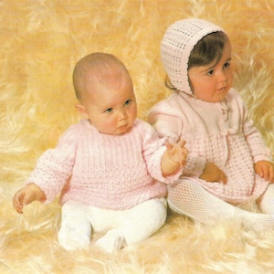 May include: Two babies wearing pink knitted outfits. One baby is wearing a pink knitted sweater and white pants. The other baby is wearing a pink knitted dress and white tights. Both babies are wearing pink knitted hats.