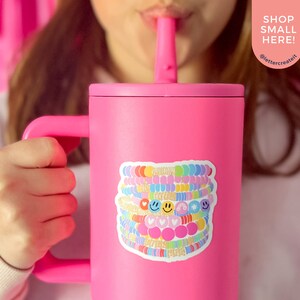 May include: Pink stainless steel travel mug with a handle and a colorful sticker of stacked bracelets with smiley faces, hearts, and stars. The sticker says "Shop Small Here!" and "@lettercreateit".