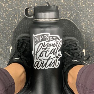 K&ouml;nnte beinhalten: Eine schwarze Wasserflasche mit einem wei&szlig;en Aufkleber, auf dem "Unterst&uuml;tze deinen lokalen K&uuml;nstler" steht.