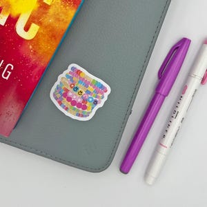 May include: A gray notebook with a sticker of colorful bracelets with smiley faces, a purple pen, and a white pen with pink accents. A book with the text "LIVING FAR" is also visible.