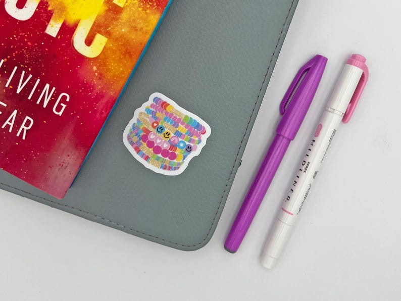 May include: A gray notebook with a sticker of colorful bracelets with smiley faces, a purple pen, and a white pen with pink accents. A book with the text "LIVING FAR" is also visible.