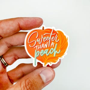 May include: A die-cut sticker with an orange pumpkin design and the phrase "Sweeter Than a Peach" in white and teal script. The sticker is held in a hand against a white background.