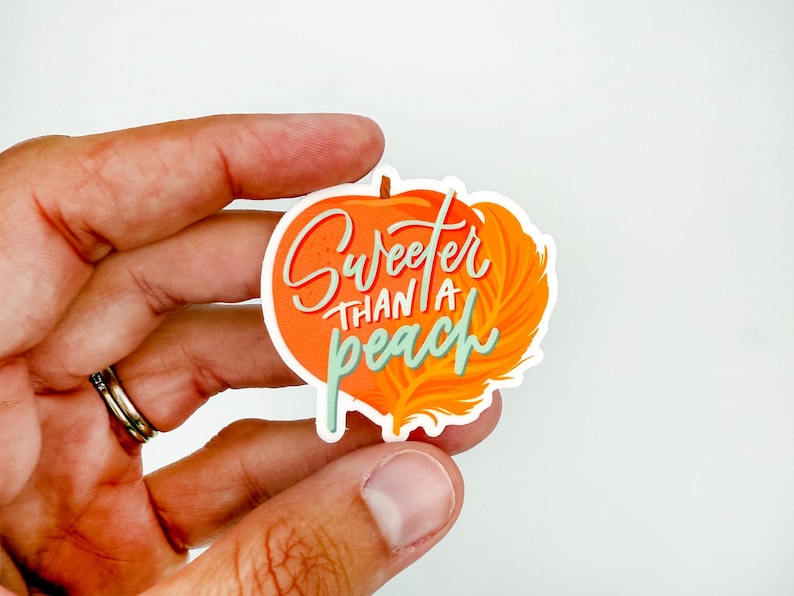 May include: A die-cut sticker with an orange pumpkin design and the phrase "Sweeter Than a Peach" in white and teal script. The sticker is held in a hand against a white background.
