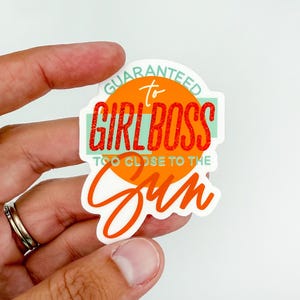 May include: A white sticker with the text "GUARANTEED to GIRL BOSS TOO CLOSE TO THE SUN" in orange, red, and teal. The sticker is held by a hand with a silver ring.