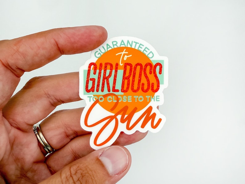 May include: A white sticker with the text "GUARANTEED to GIRL BOSS TOO CLOSE TO THE SUN" in orange, red, and teal. The sticker is held by a hand with a silver ring.