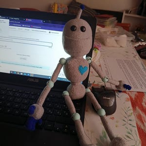 May include: A handmade gray robot doll with a blue heart and blue accents. The doll has a rounded head with two black eyes and a blue antenna. The arms and legs are jointed. The doll is sitting on a laptop.