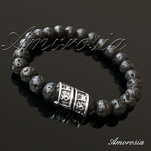 Puede incluir: Pulsera de cuentas de roca volcánica negra con un dije plateado con un patrón repetido de runas. La pulsera está sobre un fondo negro.