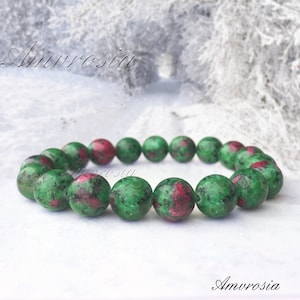 Puede incluir: Un brazalete de piedras preciosas verdes y rosas con cuentas redondas. El brazalete está hecho de piedras naturales y tiene un aspecto natural y rústico. El brazalete está sobre un fondo blanco.
