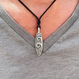 Könnte beinhalten: Eine silberne Halskette mit einem schwarzen Band. Der Anhänger ist in Form eines Surfbretts mit einem Wellendesign.