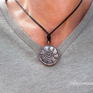 Könnte beinhalten: Eine silberne Halskette mit einem runden Anhänger, der ein Spiralmuster und Sonnenstrahlen zeigt. Der Anhänger ist an einer schwarzen Schnur befestigt.