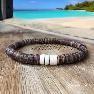 May include: A brown coconut shell beaded bracelet with white coconut shell beads. The bracelet is on a wooden surface with a blurred background of a beach.