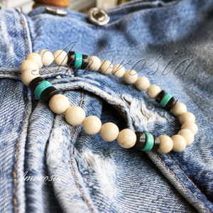 May include: A white beaded bracelet with turquoise and black accents. The bracelet is on a blue denim background.