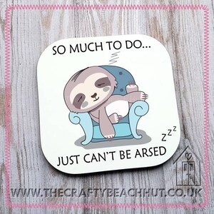 May include: A square coaster featuring a cartoon sloth asleep in a blue armchair. The text reads "SO MUCH TO DO... JUST CAN'T BE ARSED" in black font. The coaster has a white background and a pink stitched border.