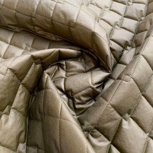 May include: Close-up of olive green quilted fabric. The fabric has a diamond pattern created by stitched lines. The material is slightly shiny and has a soft, pliable appearance. The fabric is folded, showing the texture and depth of the material.
