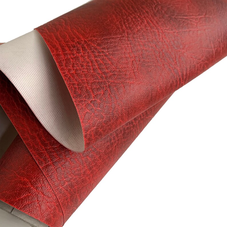 May include: Close-up of a red faux leather fabric with a textured surface. The fabric is folded over, revealing a white backing.