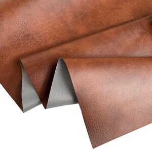 May include: Close-up of a brown faux leather fabric with a textured surface. The fabric is folded in thirds, revealing a gray backing.