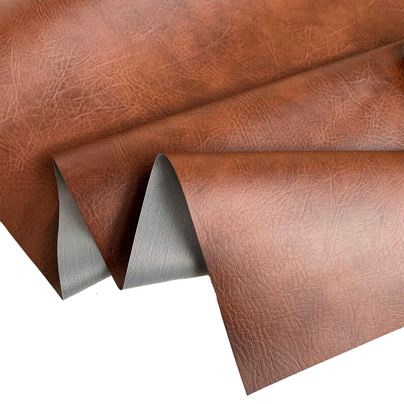 May include: Close-up of a brown faux leather fabric with a textured surface. The fabric is folded in thirds, revealing a gray backing.