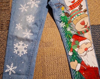 May include: Light blue denim jeans featuring hand-painted holiday designs. The left leg displays white snowflakes, while the right leg showcases a snowman, a reindeer, and Santa Claus alongside a green Christmas tree. A pink tag labeled 10 is visible.