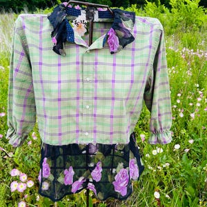 May include: A green and purple plaid shirt with a floral design on the collar and hem. The shirt has long sleeves with ruffled cuffs. The bottom hem is a black mesh with purple roses.