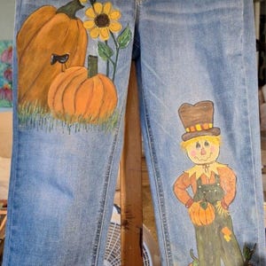 May include: Light blue denim jeans featuring hand-painted autumn designs. The left leg displays orange pumpkins, yellow sunflowers, and a small black bird. The right leg shows a scarecrow in green overalls. The cuffs feature a red wagon with pumpkins and the text Happy Fall Y'all.